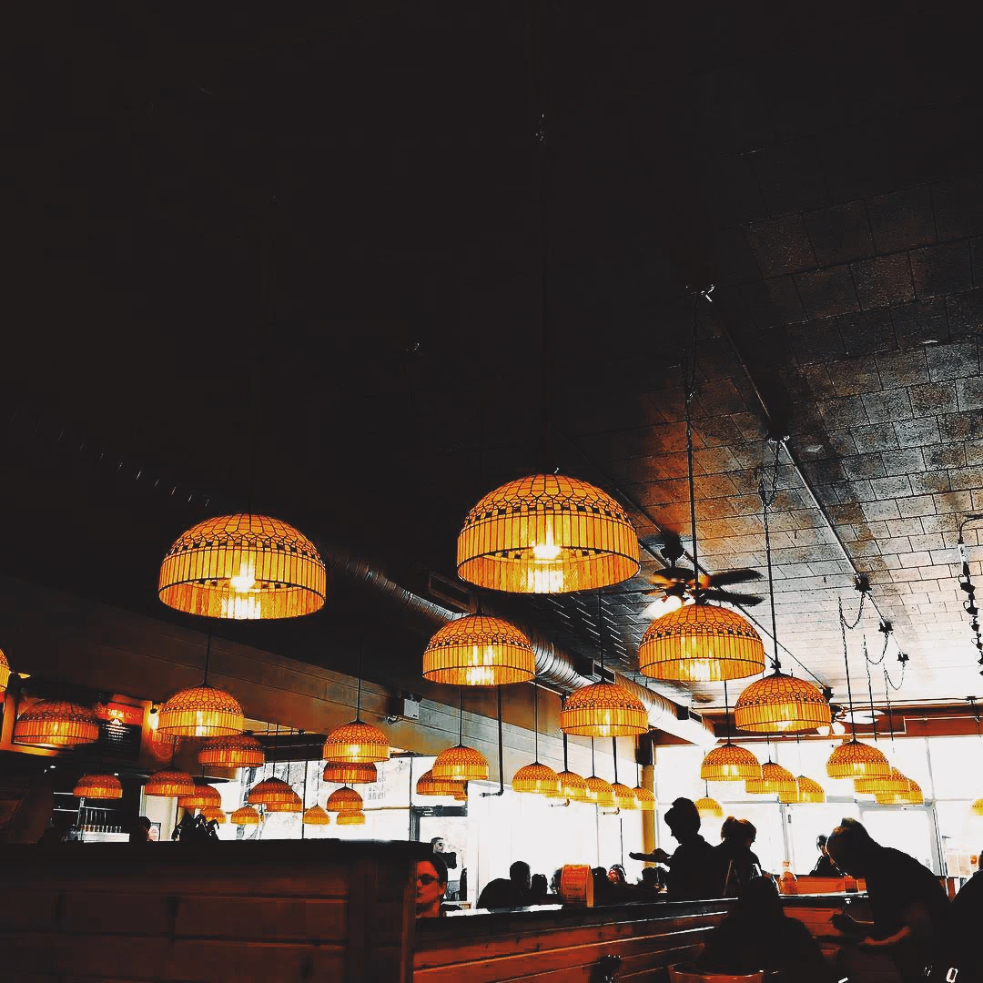 Rows of warm wicker pendant lamps hanging from a dark restaurant ceiling above the silhouettes of diners