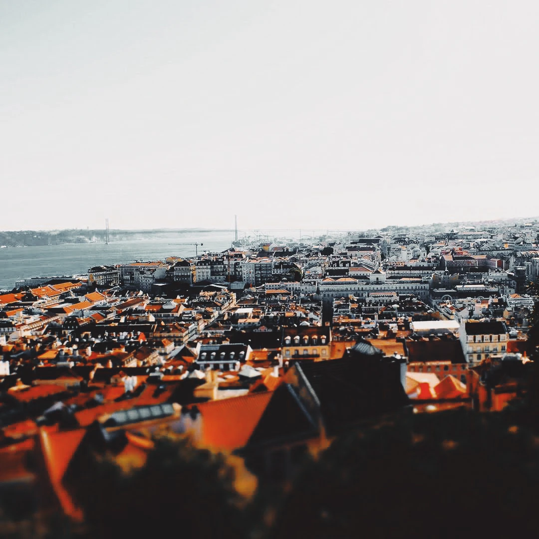 Tilt-shift aerial view of Lisbon's terracotta rooftops stretching toward the Tagus river and the 25 de Abril bridge