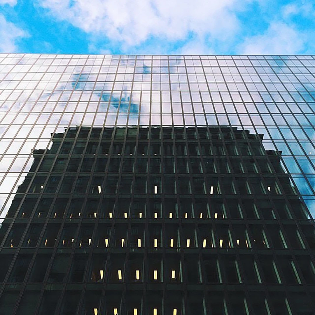 Glass-clad office tower reflecting a cloud-filled blue sky, with an older dark building visible through the facade