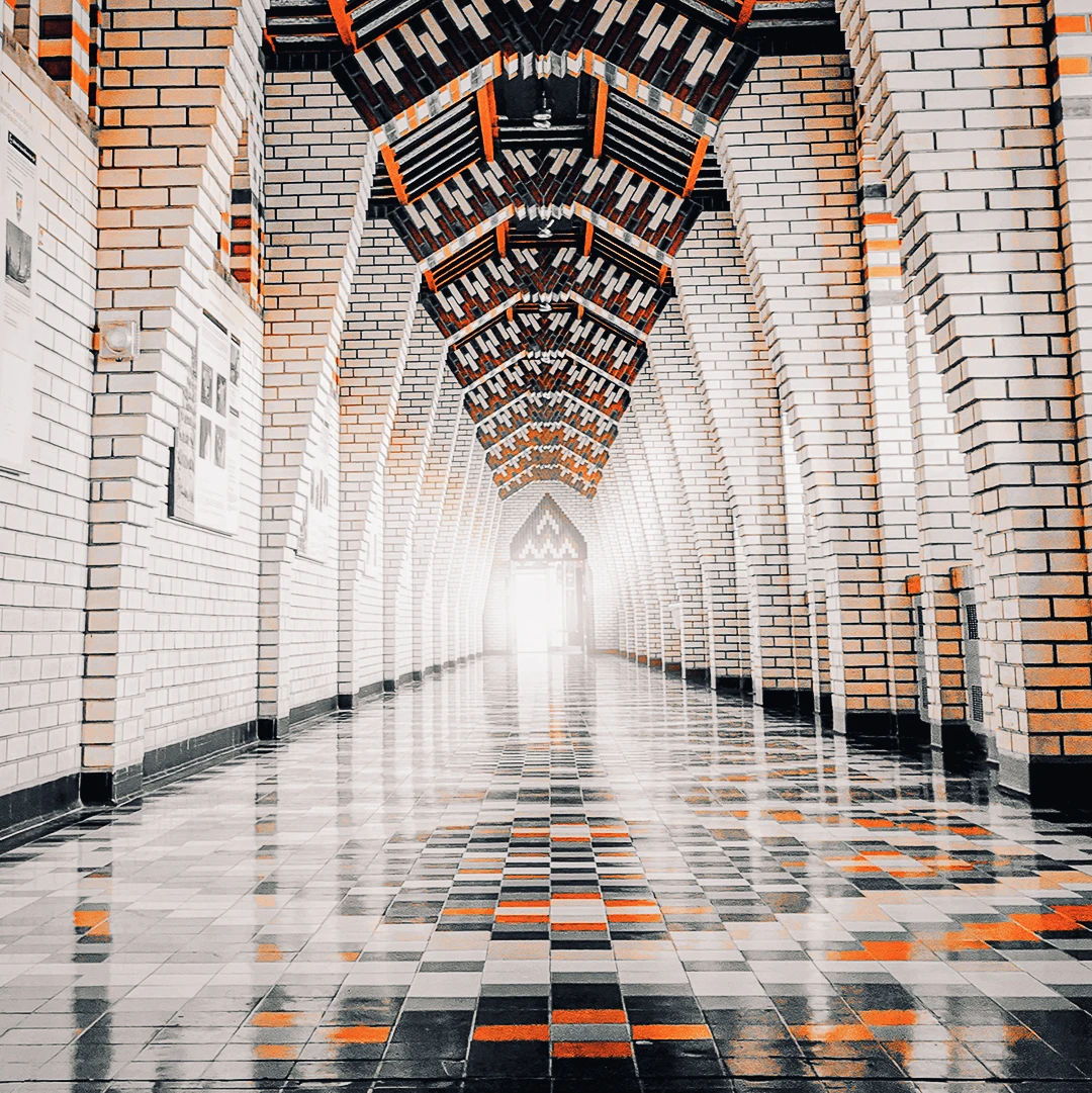 A long symmetrical corridor with ornate brick arches, patterned ceiling, and a chequered floor leading toward a bright exit