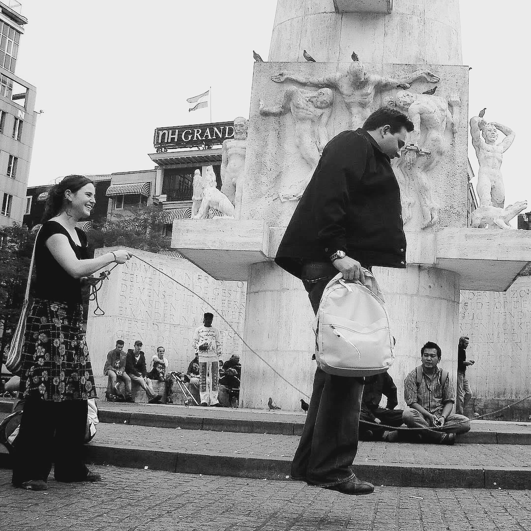 Black and white street scene in Mumbai with pedestrians moving past a monument, a seated musician visible in the background
