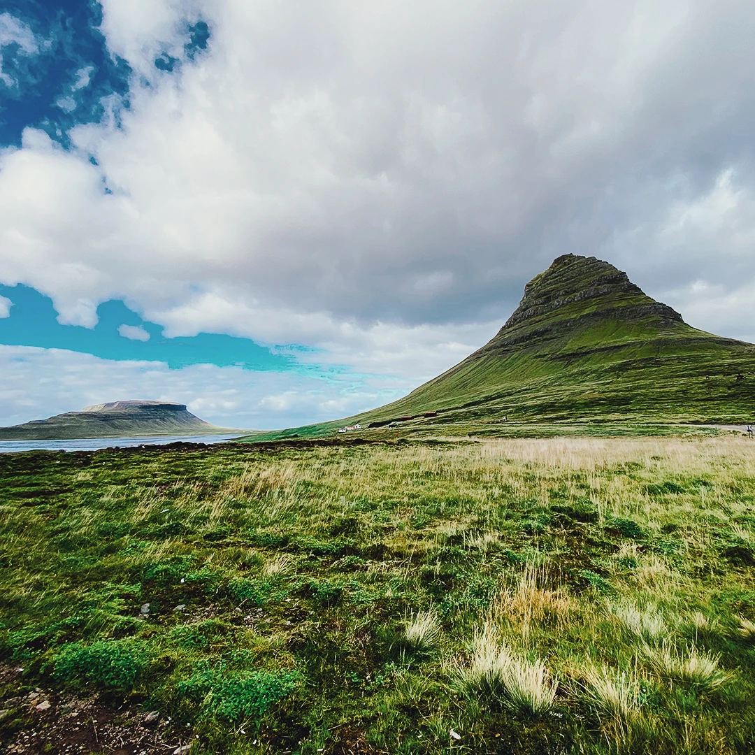 The iconic cone-shaped Kirkjufell mountain rising from green Icelandic grassland under a dramatic cloudy sky