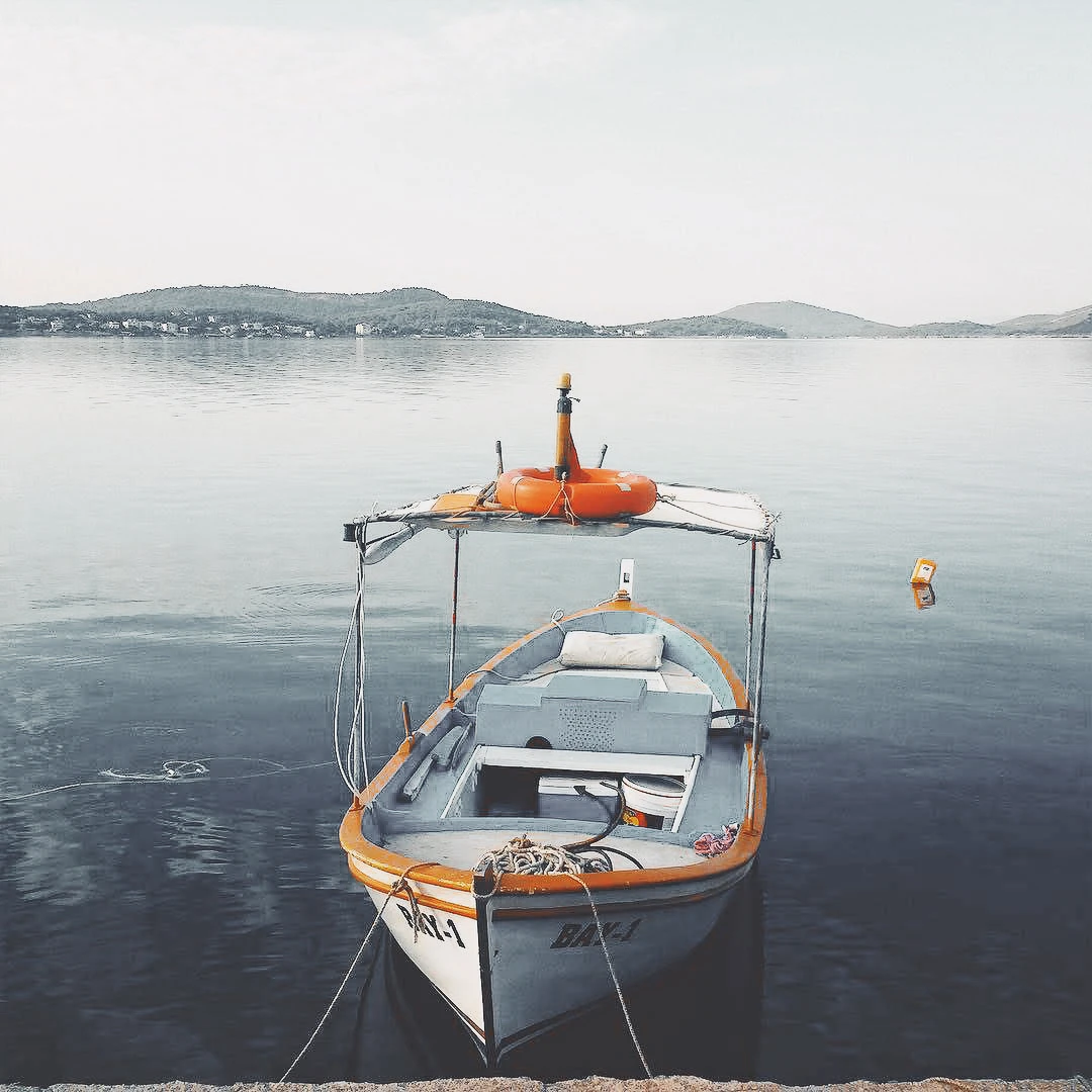 A small wooden fishing boat with orange life rings moored at a calm Adriatic bay, hills visible in the hazy distance