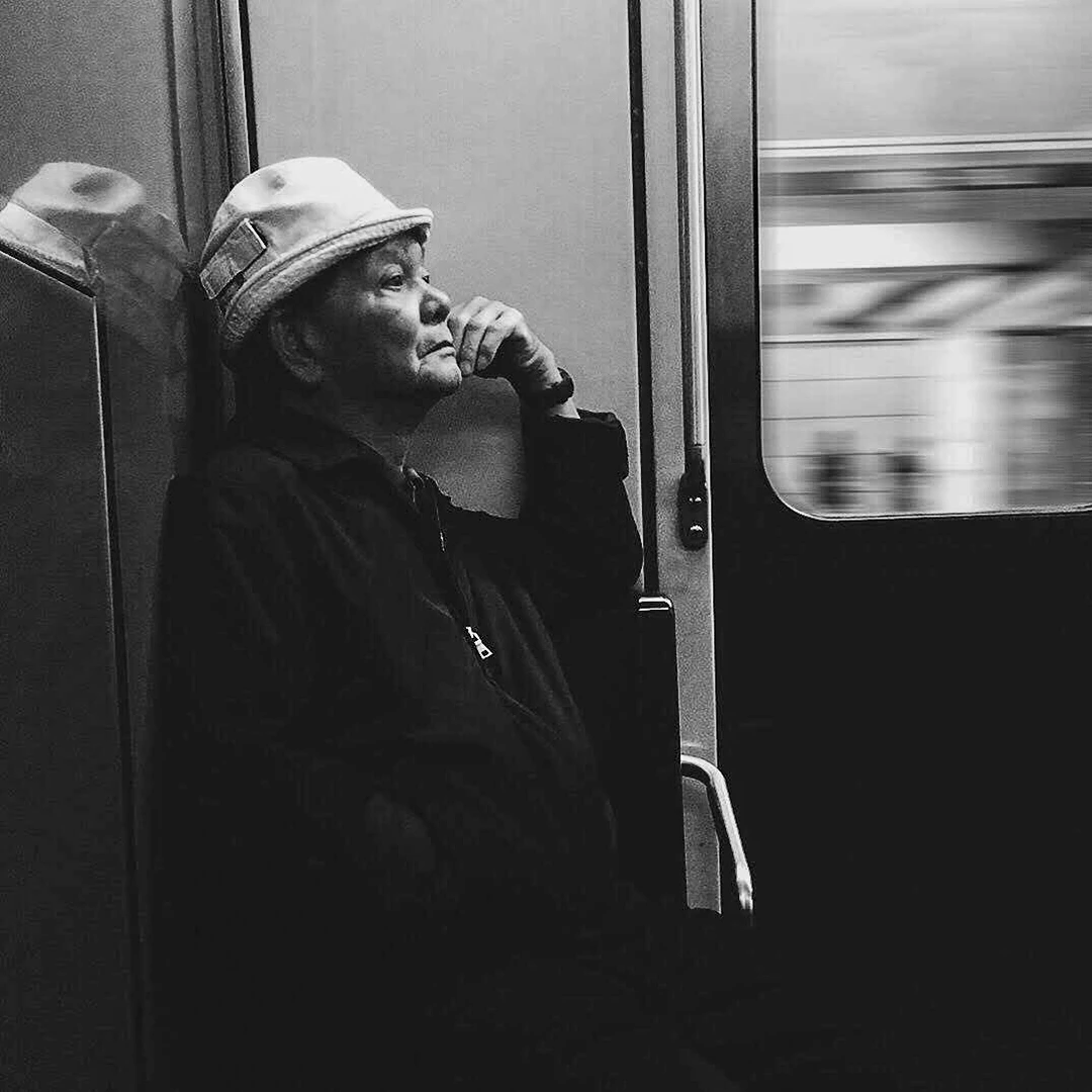 Black and white portrait of an older man in a flat cap leaning against a subway car door, lost in thought as the platform blurs past