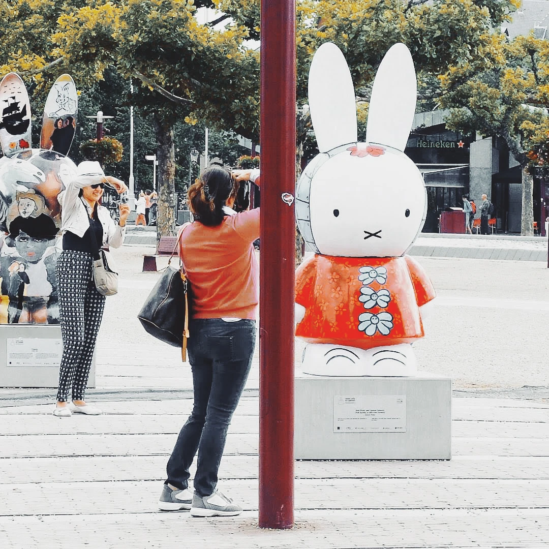 A woman photographing a large Miffy bunny sculpture in an orange floral dress on a sunny city square
