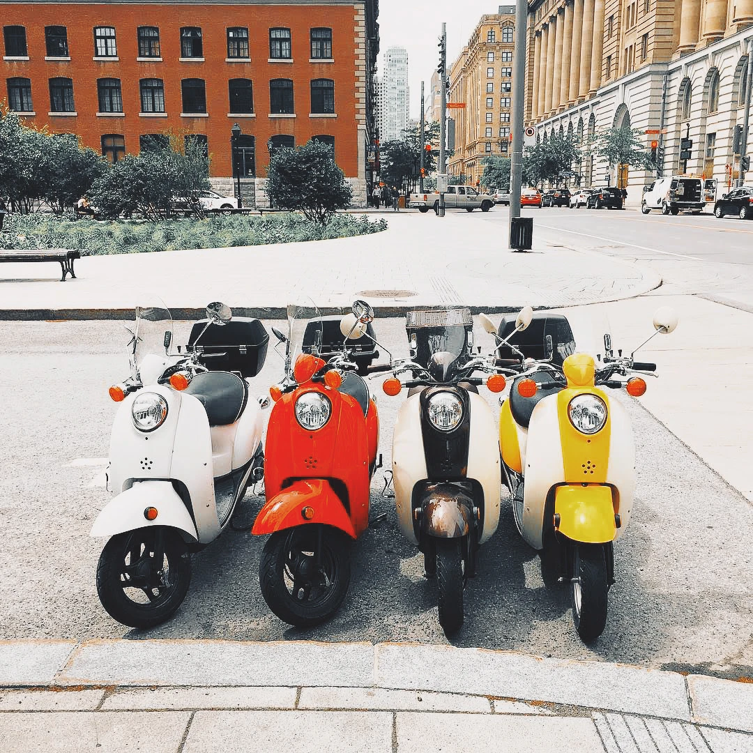 Four colourful scooters — white, red, white, and yellow — parked in a row on a city sidewalk