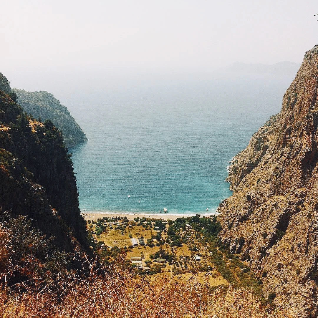 A narrow valley opening between rocky cliffs to reveal a turquoise bay and small village far below