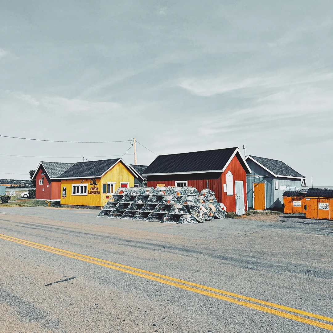 Colourful wooden fishing sheds and stacked lobster traps along a quiet coastal road under an overcast sky