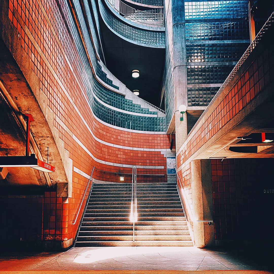 A dramatic subway staircase flanked by curved red brick walls and tiled balconies descending into a pool of light